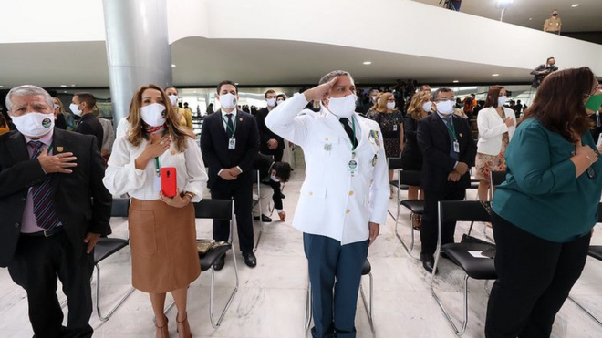 Médicos de máscara em pé, perto de cadeiras enfileiradas, um deles prestando continência