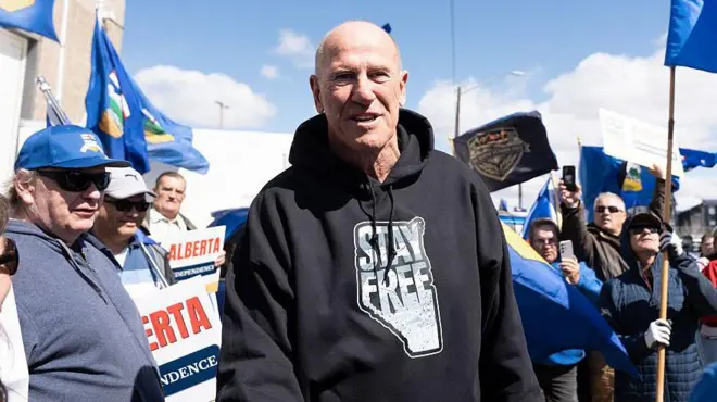 Mitch Sylvestre, director de Stay Free Alberta, un hombre de mediana edad, con poco pelo, viste una sudadera negra que lee "Queda libre" sobre el mapa de la provincia de Alberta. Está rodeado de simpatizantes que ondean banderas de Alberta