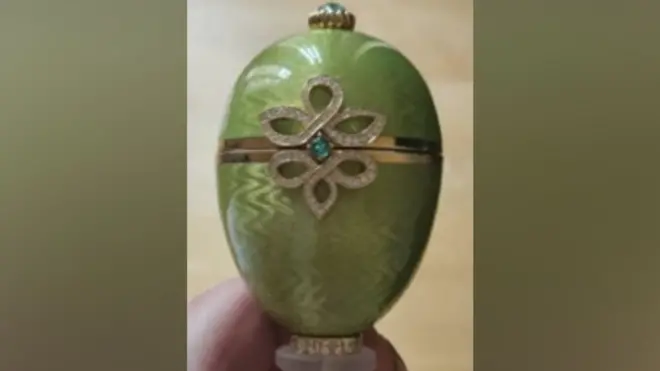 Zbliżenie zielonej ozdoby w kształcie jajka Fabergé ze złotą opaską pośrodku i dekoracyjnym wzorem w kształcie diamentu z małym niebiesko-zielonym kamieniem szlachetnym.