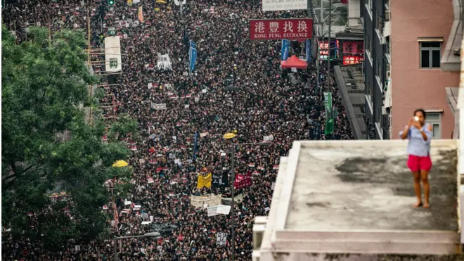 A woman stands on a rooftop taking a photo of the crowd below in this artistic composition