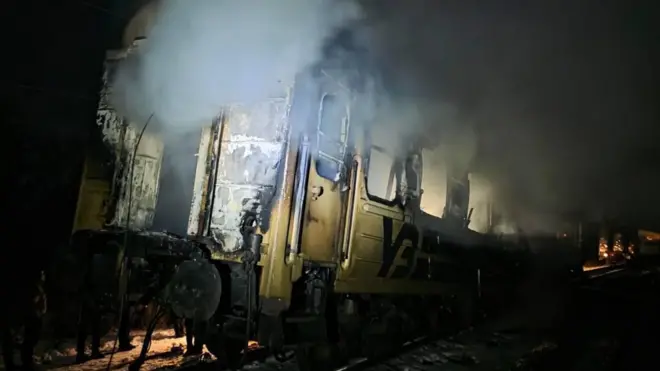 Обгорілий біло-синій вагон із написом "УЗ": його стінки почорнілі й обвуглені, частини обшивки зірвані, всередині видно сліди вогню. З даху й вікон підіймається густий білий дим. Освітлення різке, ніби від ліхтарів рятувальників чи прожекторів. Навколо темрява, а далі в глибині видно інші вагони та відблиски вогню.