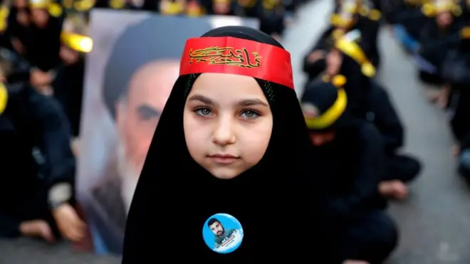 Bé gái Lebanon mặc đồ đen, phía sau là hình cố Giáo chủ đạo Hồi phái Shia của Iran trong một cuộc tuần hành ủng hộ lực lượng vũ trang Hezbollah. Tehran bị cho là có tham vọng lập Vành đai hình trăng lưỡi liềm Shia cắt ngang Trung Đông
