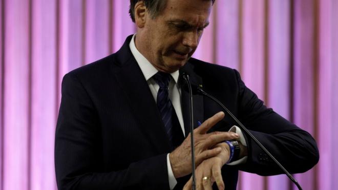 Bolsonaro olha para relógio no pulso durante evento, perto de microfone