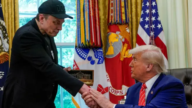 Сидящий Трамп держит двумя руками руку стоящего Маска, в другой руке Маск держит коробку с золотым ключом