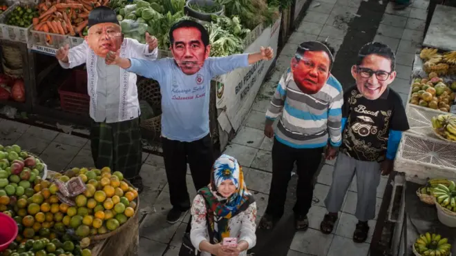 Seorang perempuan berfoto bersama warga yang mengenakan topeng pasangan Capres-Cawapres Joko Widodo-Ma'ruf Amin dan Prabowo Subianto-Sandiaga Uno dalam aksi di Pasar Gede, Solo, Jawa Tengah, Jumat (10/8). Aksi tersebut berharap pilpres 2019 berjalan dengan aman dan damai.