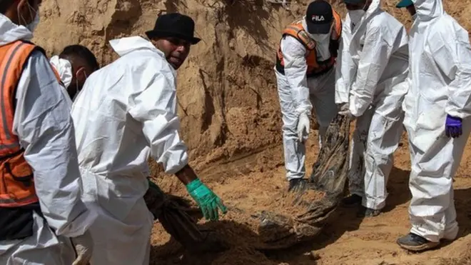 Men wearing white boiler suits drag a body wrapped in cloth from the ground on 21 April 2024