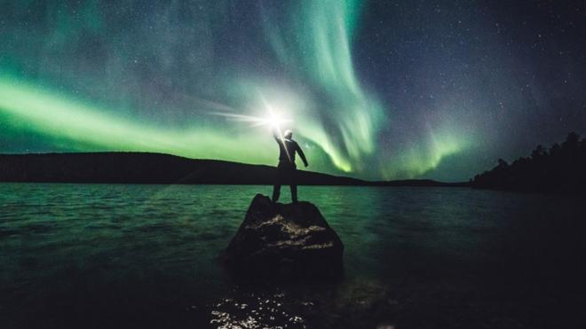 Homem ergue lanterna, com luzes da aurora boreal ao fundo