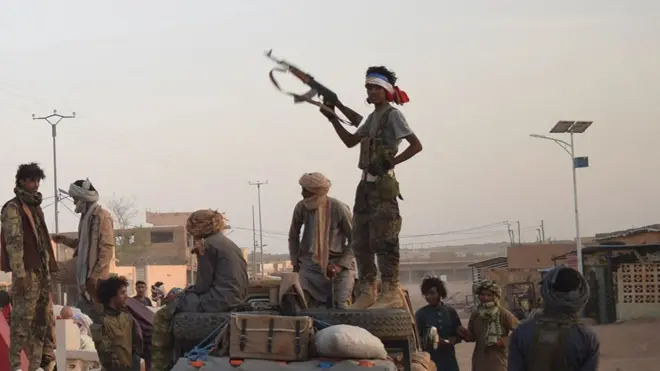 A group of fighters in camouglage stand around a pick-up truck on a dusty road. One man in near silhouette - standing on the vehicle - is holding an automatic rifle up. On the left of the picture is the brown and yellow legs of a sculpture.