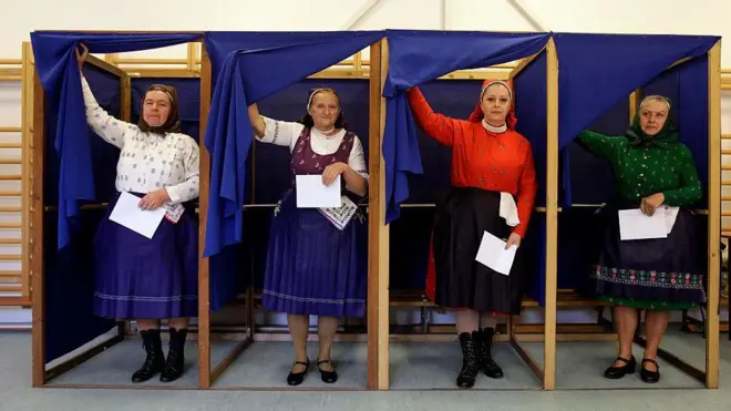 geleneksel Macar kıyafetleri giymiş kadınlar aynı anda oy kabininden çıkıyor. Hepsi renkli etekler ve bluzlar giymiş. Yaşları ileri görünüyor.