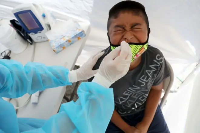 A young boy wearing a luminous face mask is shown from above pulling a face as he gets a Covid-19 test, the healthcare worker doing the swab is wearing blue PPE and gloves