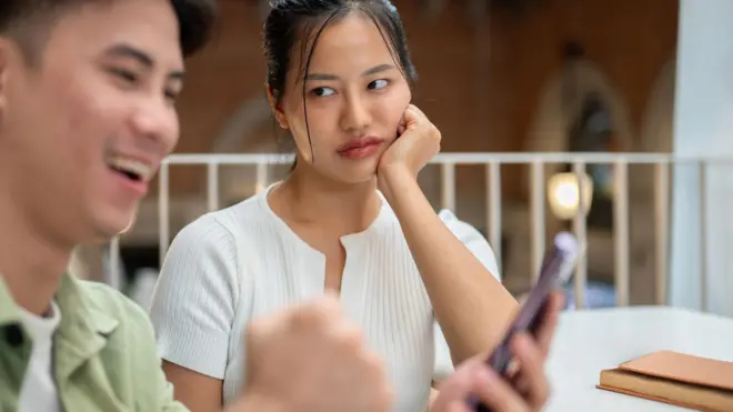 An unhappy Asian woman is feeling jealous, looking at her boyfriend while he is texting on his phone 