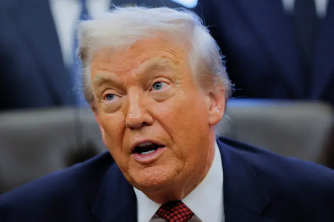 Близький портрет Трампа у темному діловому костюмі з білою сорочкою та червоно-чорною краваткою у клітинку. У тлі видно розмиті фігури інших людей у костюмах, що стоять позаду.