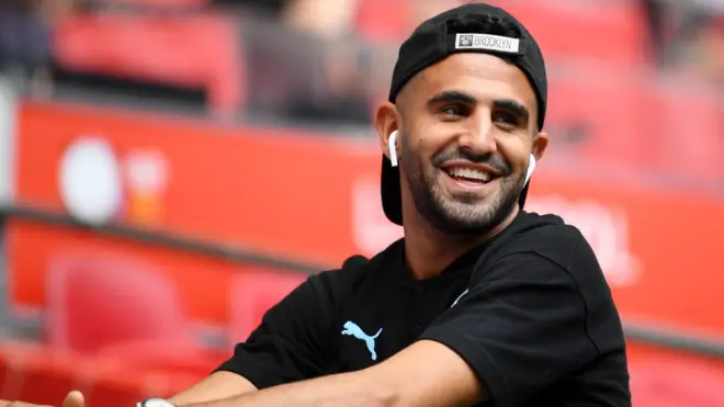 A picture of Riyad Mahrez at Wembley, smiling, with headphones in and wearing a cap backwards