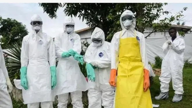 Awọn oṣiṣẹ eleto ilera dira delẹ lati dena ikọlu Ebola, aṣo funfun ni wọn wọ, ọkan ninu wọn wọ agbekọ alawọ pupa rẹsurẹsu le ori aṣọ rẹ. Marun-un ninu wọn lo han kedere ninu aworan yii, ọwọ ẹnikẹfa yọ ṣugbọn oju re ko han. Igi wa lẹyin wọn nibi ti wọn ti ya aworan yii, a si ri ile iyasọtọ ti won n ko awọn ti Ebola ba kọlu si pẹlu.