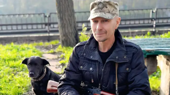 Чоловік із рушницею сидить біля собаки. 