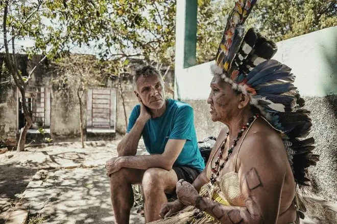 Dom e pajé sentados em área externa de casa; ela fala enquanto ele escuta