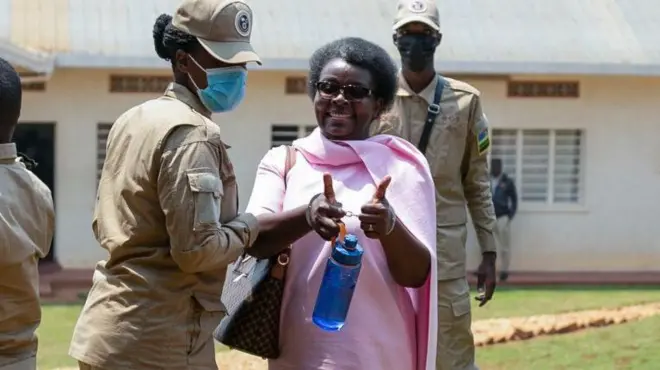 Victoire Ingabire ku rukiko muri Kanama (8) mu mwaka wa 2025. Arahagaze, amwenyura, akoze ikimenyetso cy'ibikumwe by'ibiganza bye, afashe icupa ry'ubururu ry'amazi, yambaye umwambaro w'iroza uranga imfungwa mu Rwanda. Iruhande rwe hari umucungagereza w'umugore wambaye ingofero n'imyambaro ya khaki ibaranga, hamwe n'agapfukamunwa k'ubururu bwerurutse. Inyuma ya Ingabire hahagaze undi mucungagereza w'umugabo wambaye imyambaro ya khaki ibaranga, n'agapfukamunwa k'umukara. 