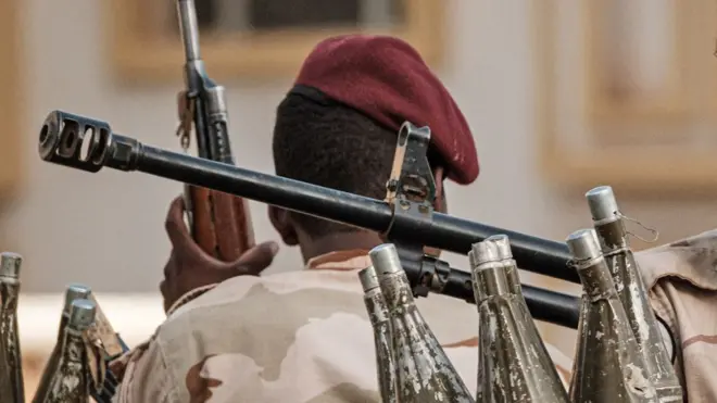 Vue de dos d'un combattant des RSF coiffé d'un béret rouge et tenant un fusil. Devant lui, on aperçoit le canon et des roquettes de mortier.