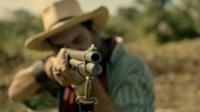 Homem branco de barba e chapéu, com rosto desfocado, segura uma espingarda e aponta para a câmera
