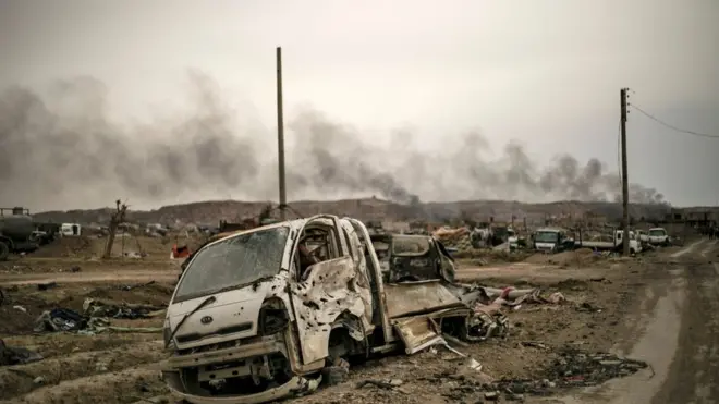 الدخان يتصاعد من خلف المركبات المدمرة والمباني المتضررة في قرية الباغوز بمحافظة دير الزور شرقي سوريا