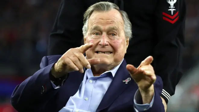 George HW Bush points to an off-camera person from his wheelchair