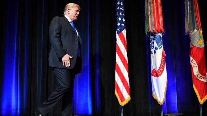 Donald Trump caminha em palco em direção ao púlpito, com bandeiras ao fundo