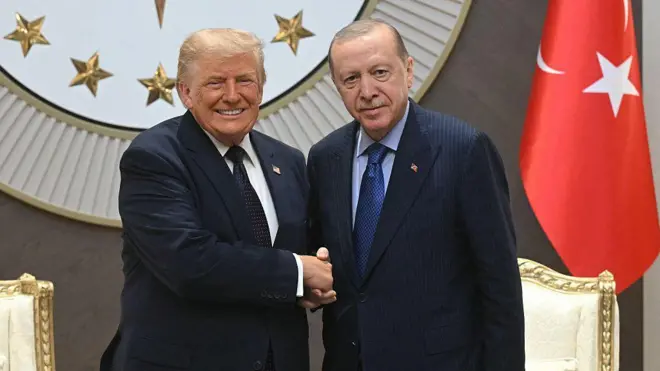Trump ve Erdoğan el sıkışırken gülümsüyorlar geride yaldızlı koltukların ve Cumhurbaşkanlığı ambleminin bölümleri görünüyor. 