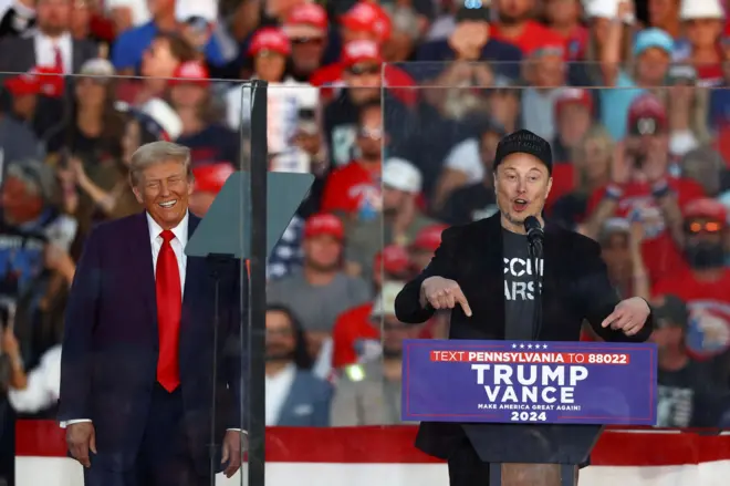 Trump sorrindo enquanto Musk discursa e gesticula; eles são observados por plateia