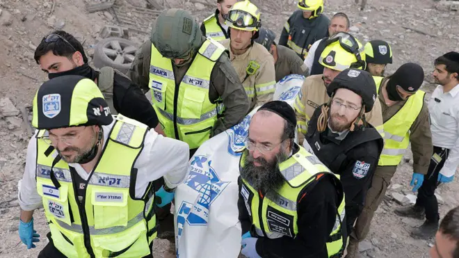 Çok sayıda İsrail'li acil durum personeli bir cesedi taşırken görülüyor. Arkada yıkıntılar görünüyor.