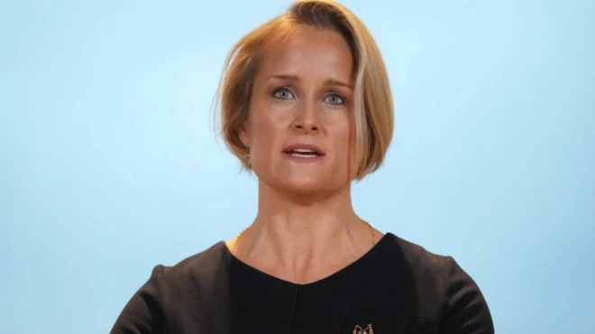 Metreweli kürsünün arkasında konuşma yapıyor. Üzerinde siyah bir elbise ve bir broş var.