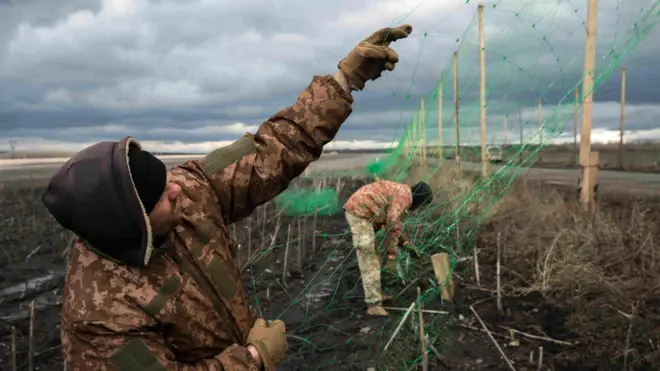 Двое украинских военных натягивают антидроновую сетку на столбах вдоль дороги 