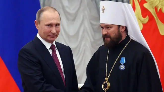Путін (ліворуч) в темному костюмі та краватці та Іларіон (праворуч) – в чорному релігійному вбранні з білим головним убором, із великим хрестом на ланцюгу на грудях і нагородою на одязі – стоять поруч у приміщенні перед великими прапорами РФ і декоративним інтер’єром.
