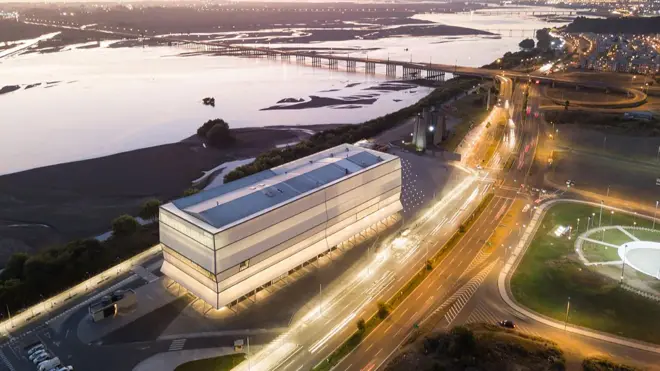 Edificio moderno junto a una carretera iluminada al atardecer, con un río amplio y puentes al fondo.