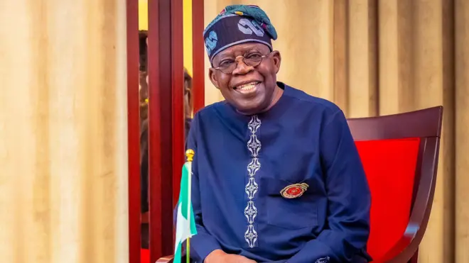 Bola Tinubu looking at di camera smiling wit Nigeria flag for im front.