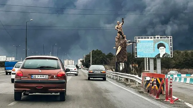 Veículos trafegam por uma rodovia, passando por uma estátua em memória aos mortos na guerra e um outdoor retratando o falecido líder supremo do Irã, o aiatolá Ali Khamenei, morto em um ataque aéreo em 28 de fevereiro, com colunas de fumaça preta subindo, em Teerã, em 8 de março.
