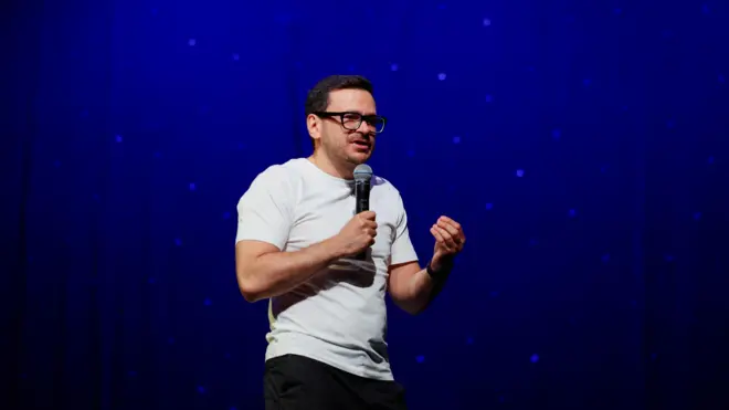 Ilja Jašin u beloj majici i sivim pantalonama na sceni drži mikrofon i priča