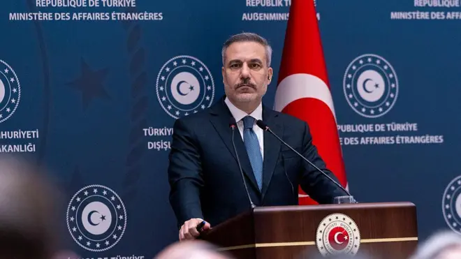 Hakan Fidan, bir kürsüde ayakta duruyor. Arkasında Türk bayrağı var ve Dışişleri Bakanlığı yazısı olan bir pano önünde duruyor.