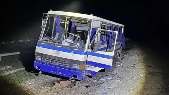 Пошкоджений синьо-білий автобус стоїть на узбіччі дороги вночі. У нього вибиті всі вікна, колес не видно – так, ніби кузов вріс у землю. Навколо видно уламки та сміття, освітлені світлом ліхтаря.