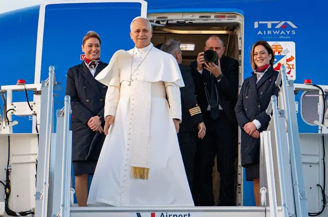 Le Pape Léon XIV dans une robe blanche à la porte d'entrée d'un avion, avec à ses côtes deux hôtesses de l'air et en arrière-plan un homme avec un appreil photo.