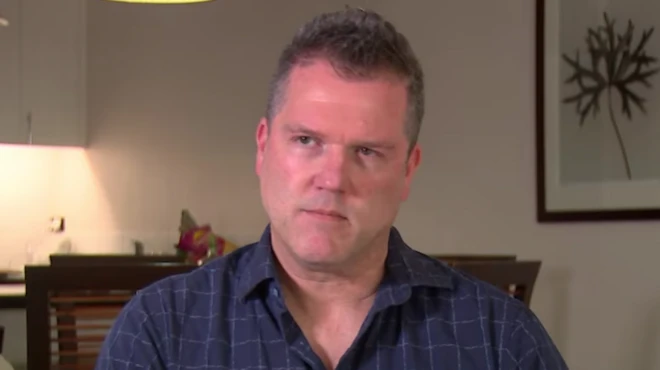 Кріс Вебстер у темно-синій картатій сорочці дивиться в камеру. Позаду нього видно обідній стіл і кухню.