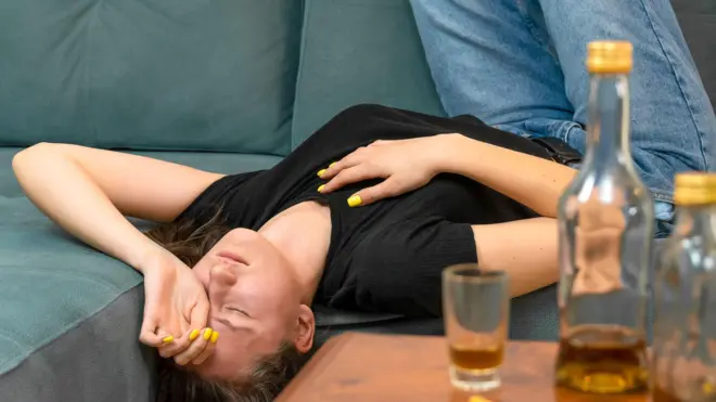 A young woman lying on a sofa upside-down with her eyes closed and hand over her face, with near-empty bottles standing on the table in front
