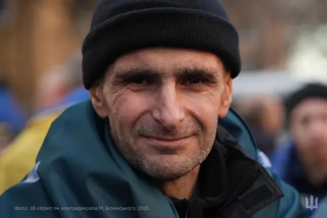 Крупний план Олександра Савова у темній вовняній шапці, що дивиться в об'єктив камери. Схудле обличчя з легкою непритістю, на вустах посмішка