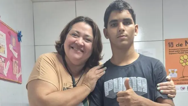 Mulher e jovem juntos em uma sala, ela sorrindo e ele fazendo sinal positivo com a mão