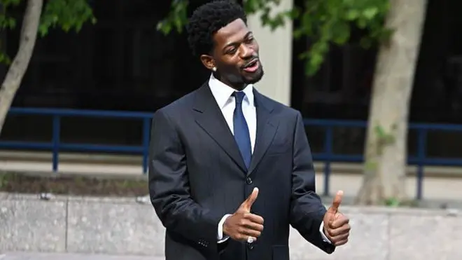 Lil Nas X делает жест большими пальцами двух рук. Он одет в темный костюм, белую рубашку и галстук