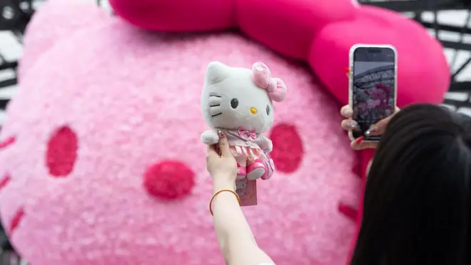 A woman holds up a Hello Kitty soft toy to take a photo with her phone. Behind the toy there is a large pink Hello Kitty head