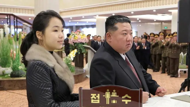Kuzey Kore lideri Kim Jong Un (sağda) ve kızı Kim Ju Ae bir otel resepsiyonunda duruyor. Arka planda lobide askerler alkışlıyor.