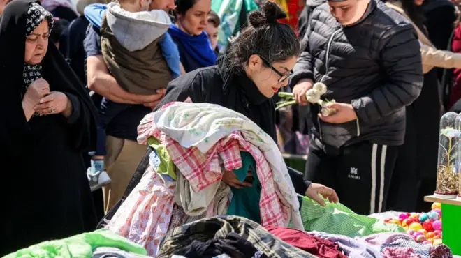 زنی در ۱۷ مارس ۲۰۲۵ در حالی که مردم برای نوروز (نوروز)، سال نو ایرانی، که امسال مصادف با ماه رمضان است، آماده می‌شوند، لباس‌های فروش را در غرفه‌ای خارج از بازار تجریش در مرکز تهران جستجو می‌کند. 