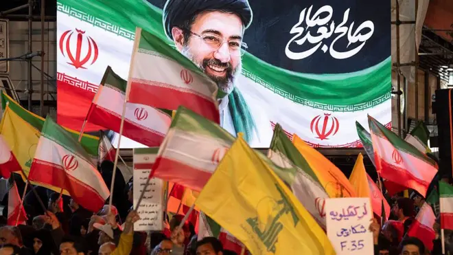 لوّح متظاهرون مؤيدون للحكومة الإيرانية بالأعلام الوطنية، وأعلام حزب الله اللبناني، ولافتة كُتب عليها بالفارسية: "صائد طائرات إف-35"، أمام صورة المرشد الأعلى الإيراني الجديد آية الله مجتبى خامنئي، خلال مراسم تشييع جثمان مجيد خادمي، رئيس جهاز المخابرات التابع للحرس الثوري الإيراني، في طهران، إيران، في 8 أبريل/نيسان 2026.