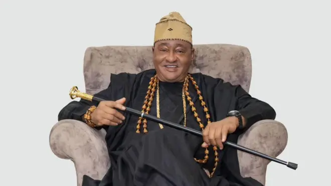 Jide Kosoko joko sori aga pẹlu ọpa lọwọ rẹ, o wọ aṣọ dudu pẹlu fila