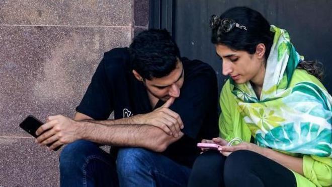 یک دختر بدون حجاب در کناری پسری نشسته و هر دو به گوشی موبایلی که دست دختر است نگاه می‌کنند